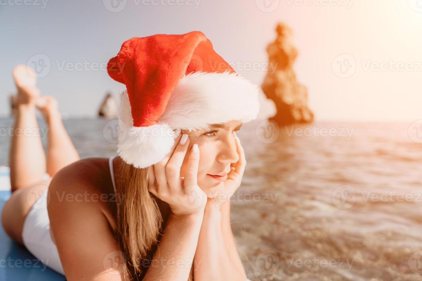 Woman sea sup. Close up portrait of happy young caucasian woman with long hair in Santa hat looking at camera and smiling. Cute woman portrait in a white bikini posing on sup board in the sea photo