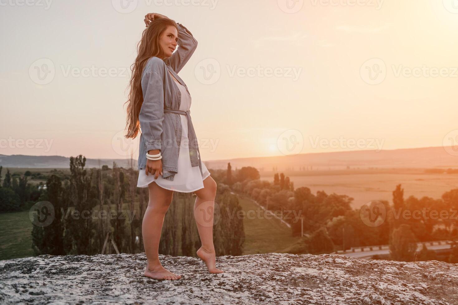 Happy woman standing with her back on the sunset in nature in summer with open hands. Romantic beautiful bride in white boho dress posing with mountains on sunset photo