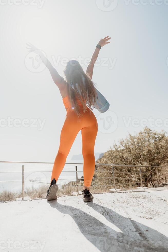 Woman in Orange Yoga Clothes with Arms Raised on a Cliff Overlooking the Ocean photo