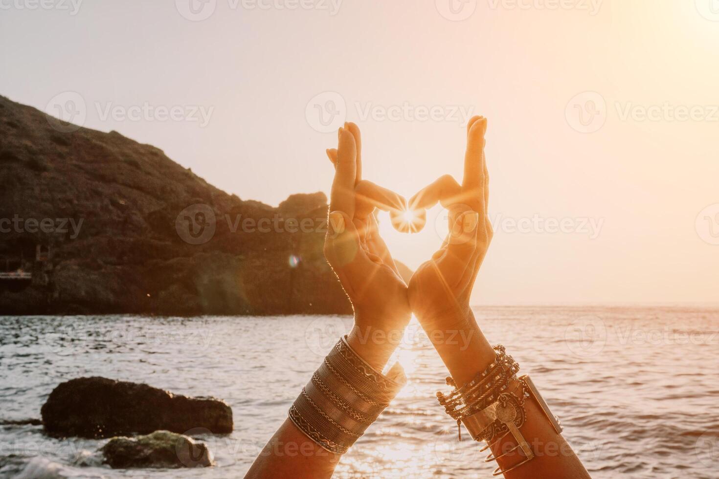 Woman sea yoga. Happy woman in a white swimsuit and boho-style bracelets making a heart shape with her hands while practicing yoga on a mat by sea at sunset. Healthy lifestyle, harmony and meditation photo