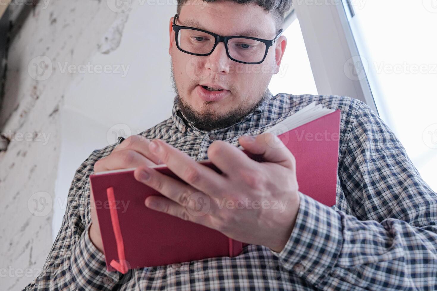 man in plaid shirt sitting at windowsill , thinking and writing in a notebook photo