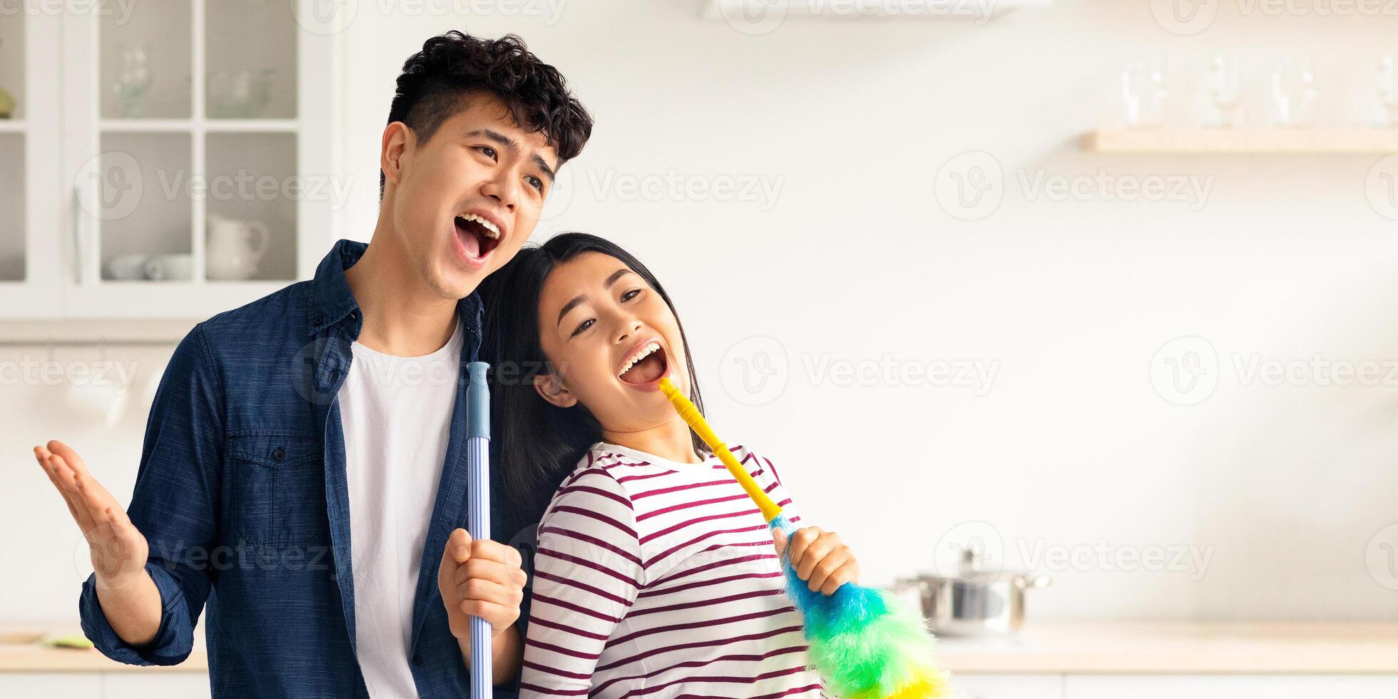 Joyful asian loving couple singing songs while cleaning kitchen, using fluffy dust brush as microphone, cheerful beautiful young man and woman imitating rock stars while house-keeping, copy space photo