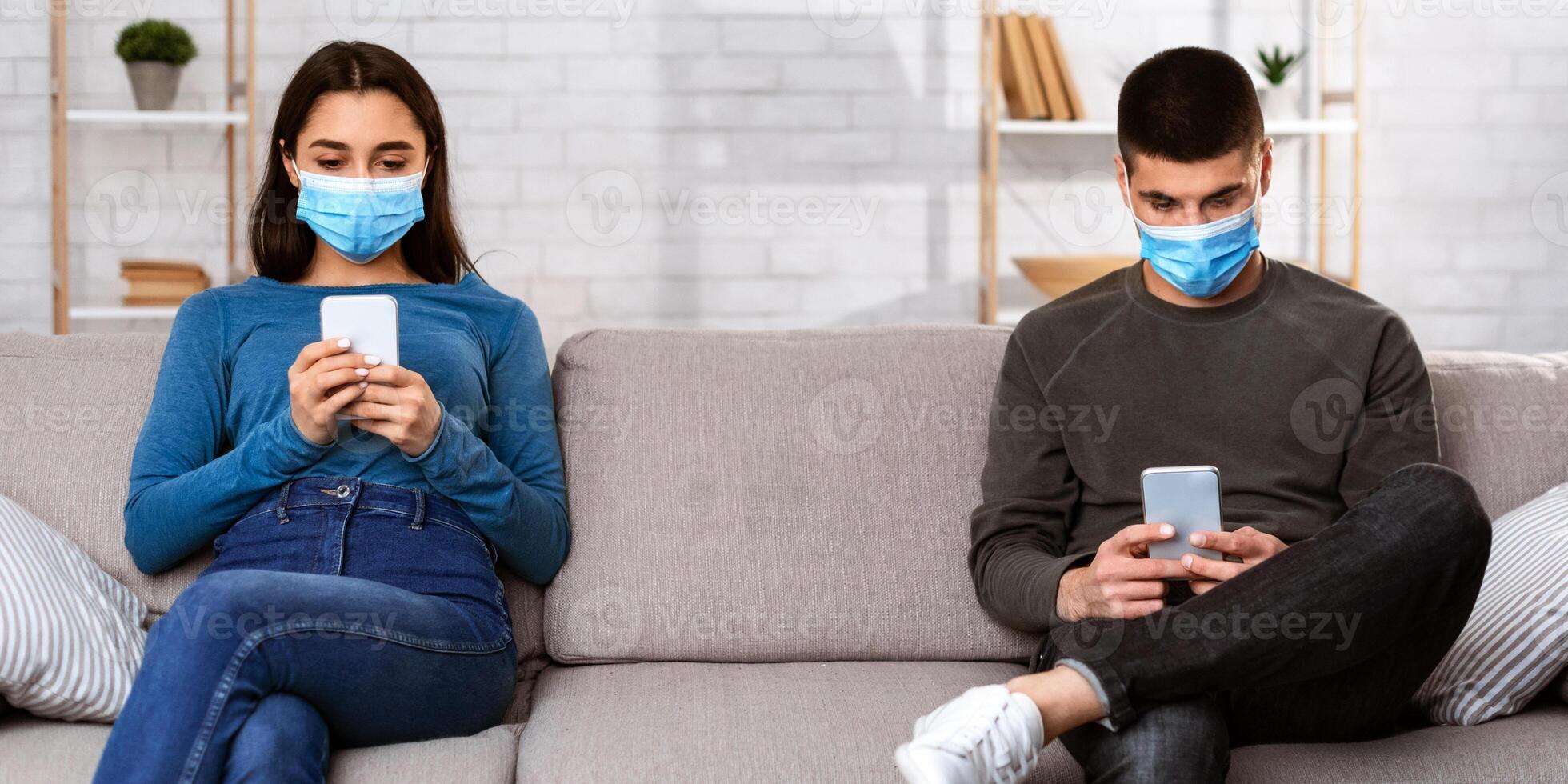 Self Isolation Concept. Young lovely couple wearing surgical face mask using smart phones, sitting on couch at home photo