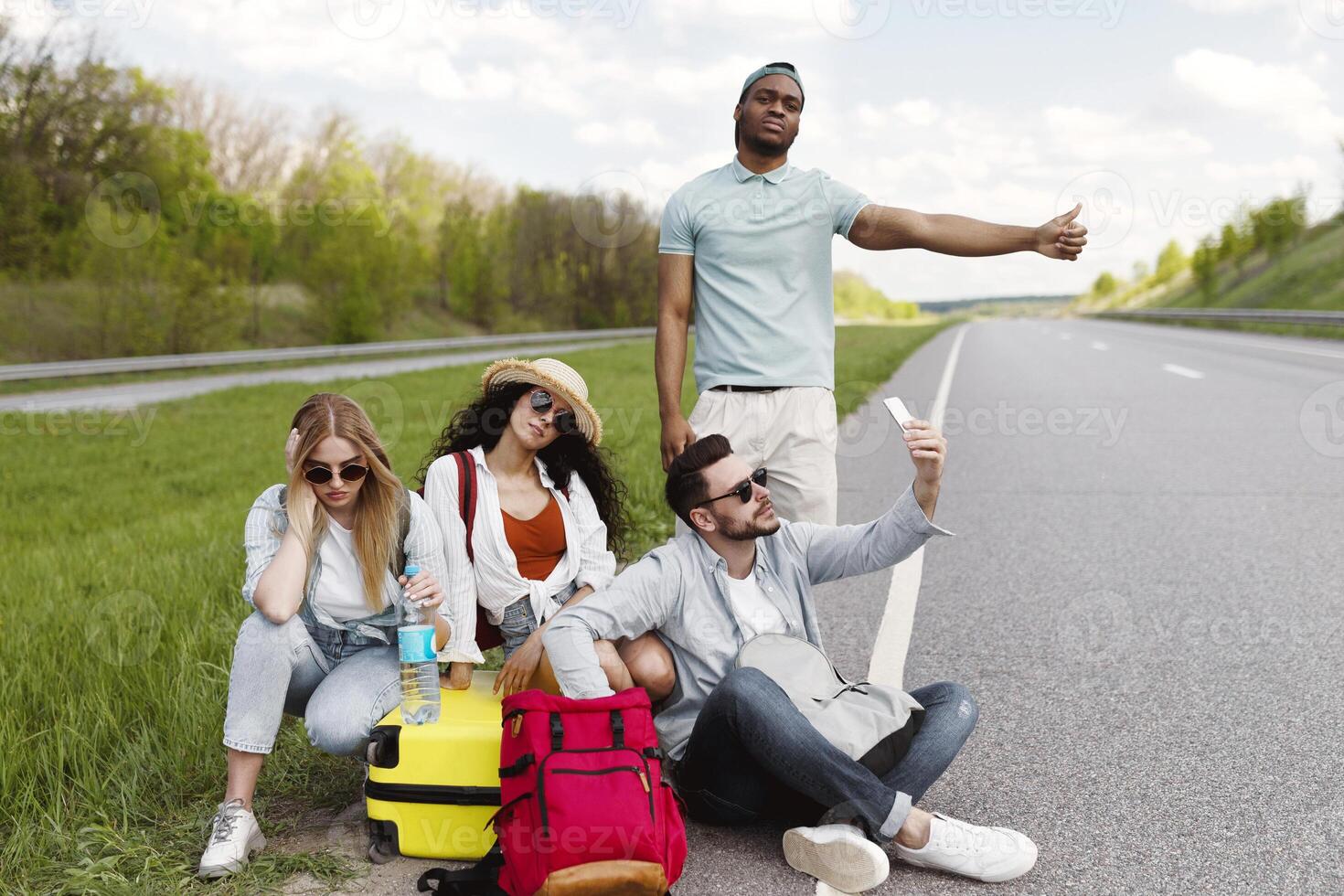 Young multiracial friends hitchhiking on highway, trying to stop car without success, feeling tired and upset on road trip, copy space. Traveling by autostop, summer vacation, tourism and friendship photo