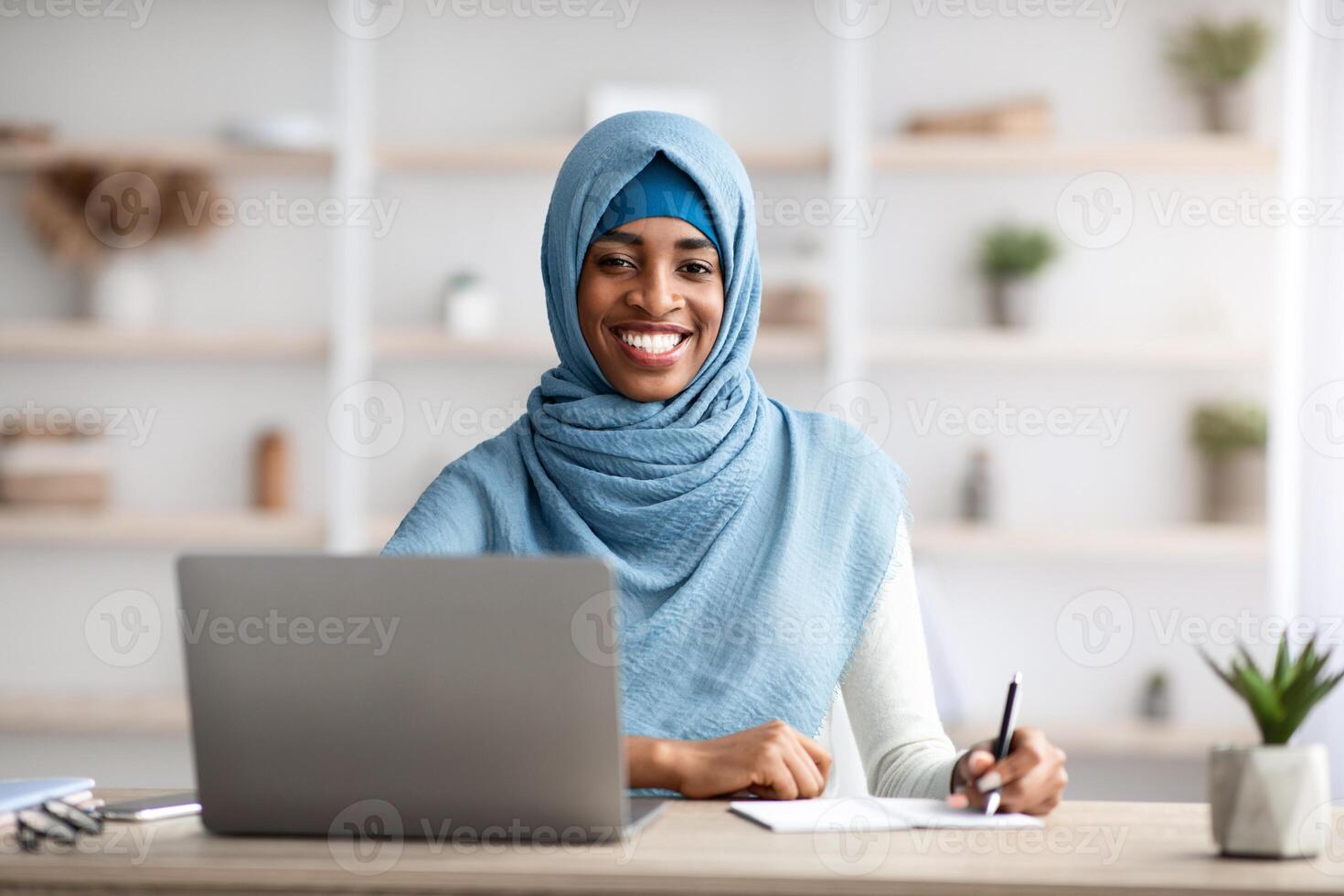 Job Opportunities For Muslim Women. Happy Black Islamic Female Sitting At Desk With Computer, Smiling Religious African Lady In Hijab Working On Laptop In modern Office And Taking Notes, Free Space photo