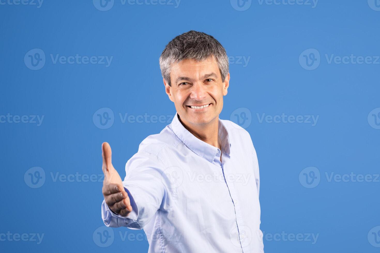 A middle aged man in his 50s stands against a solid blue background, smiling and extending his right hand in a welcoming gesture. His attire is casual, reflecting a friendly lifestyle. photo