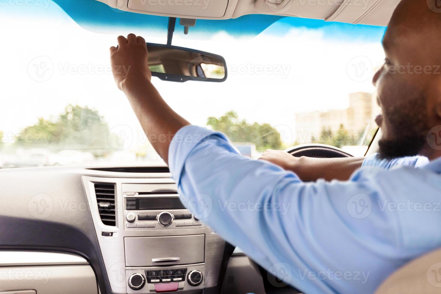 Testing Visibility. Happy Black Guy Adjusting Rear View Mirror, Enjoying New Auto, Sitting On Driver's Seat In Luxury Vehicle. Man Driving Alone, Over Shoulder View From The Back Passenger Seat photo