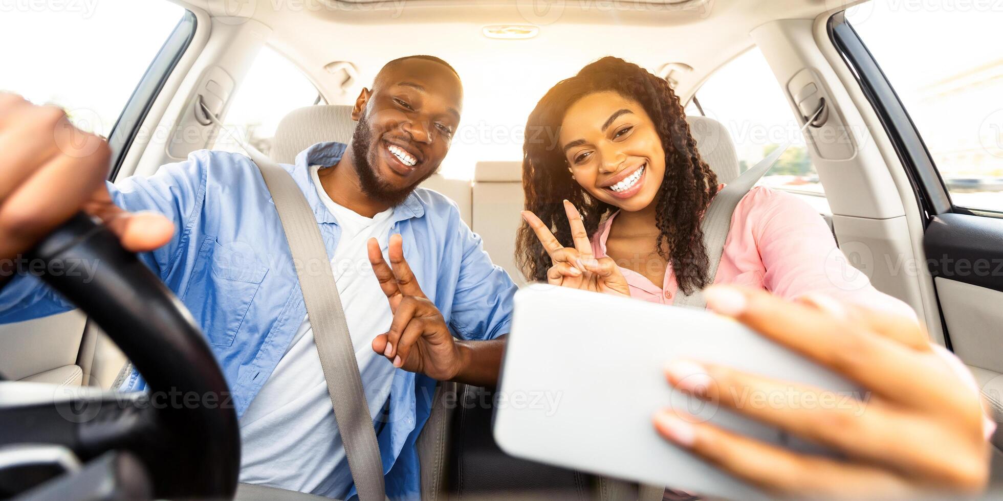 Beautiful young African American couple taking selfie inside car, using mobile phone, showing v victory peace sign gesture. Happy black man and woman going on vacation, taking self photo in auto