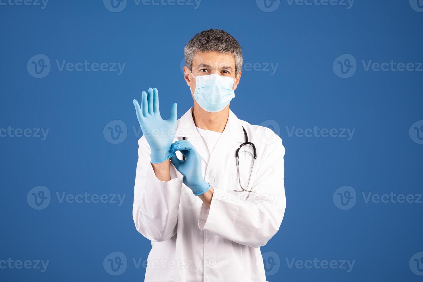 A mature Caucasian male doctor dressed in a lab coat and surgical mask is putting on blue gloves. He stands against a plain blue background, getting ready for a medical consultation. photo