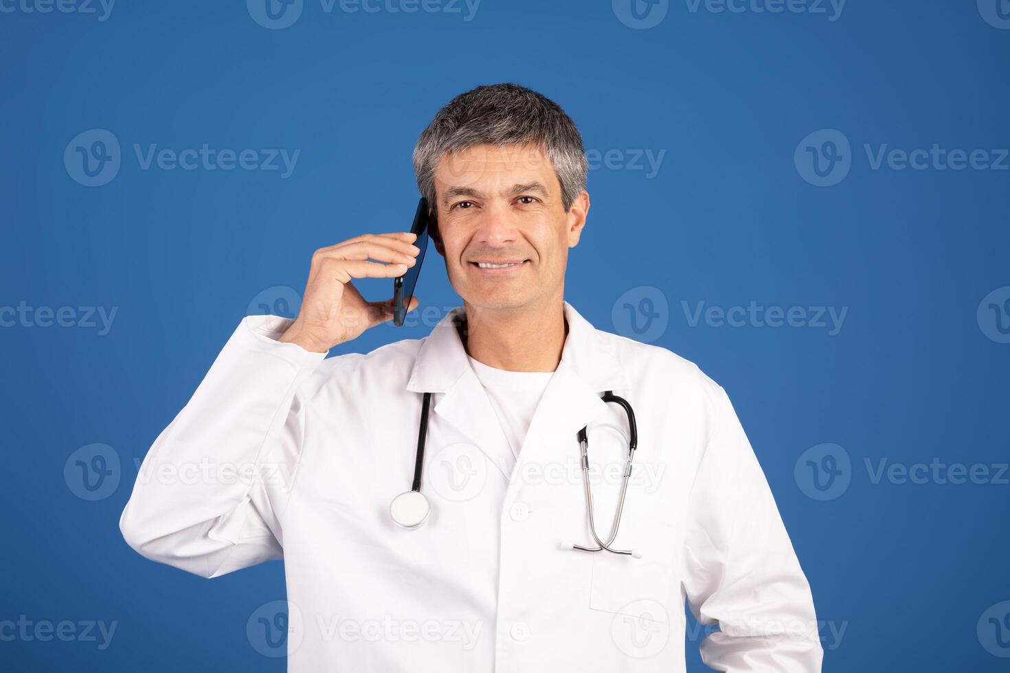 A mature Caucasian doctor in a white coat communicates on the phone, smiling. The background features a vibrant blue color, creating a lively and approachable atmosphere in a healthcare setting. photo