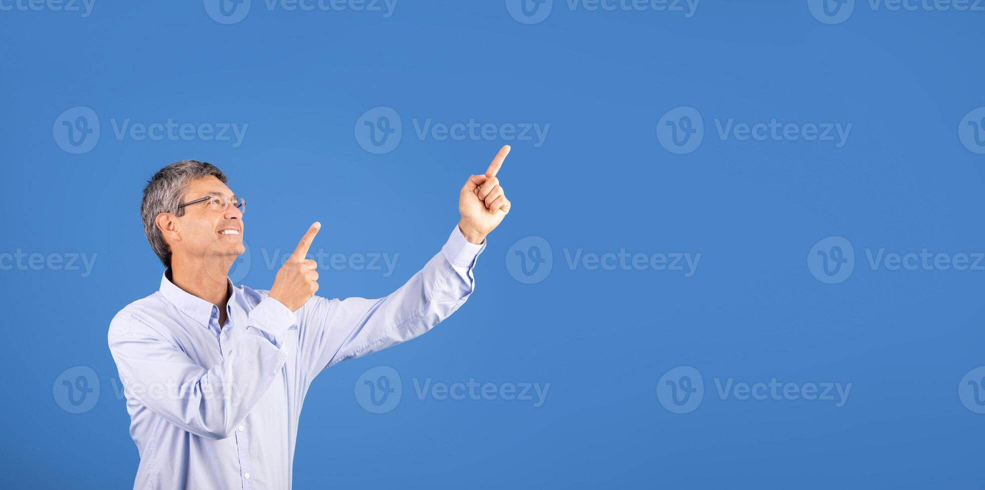A mature man in his 50s gestures with both hands, pointing upward at copy space with a big smile. The clear blue background enhances his lively expression, suggesting excitement or celebration. photo