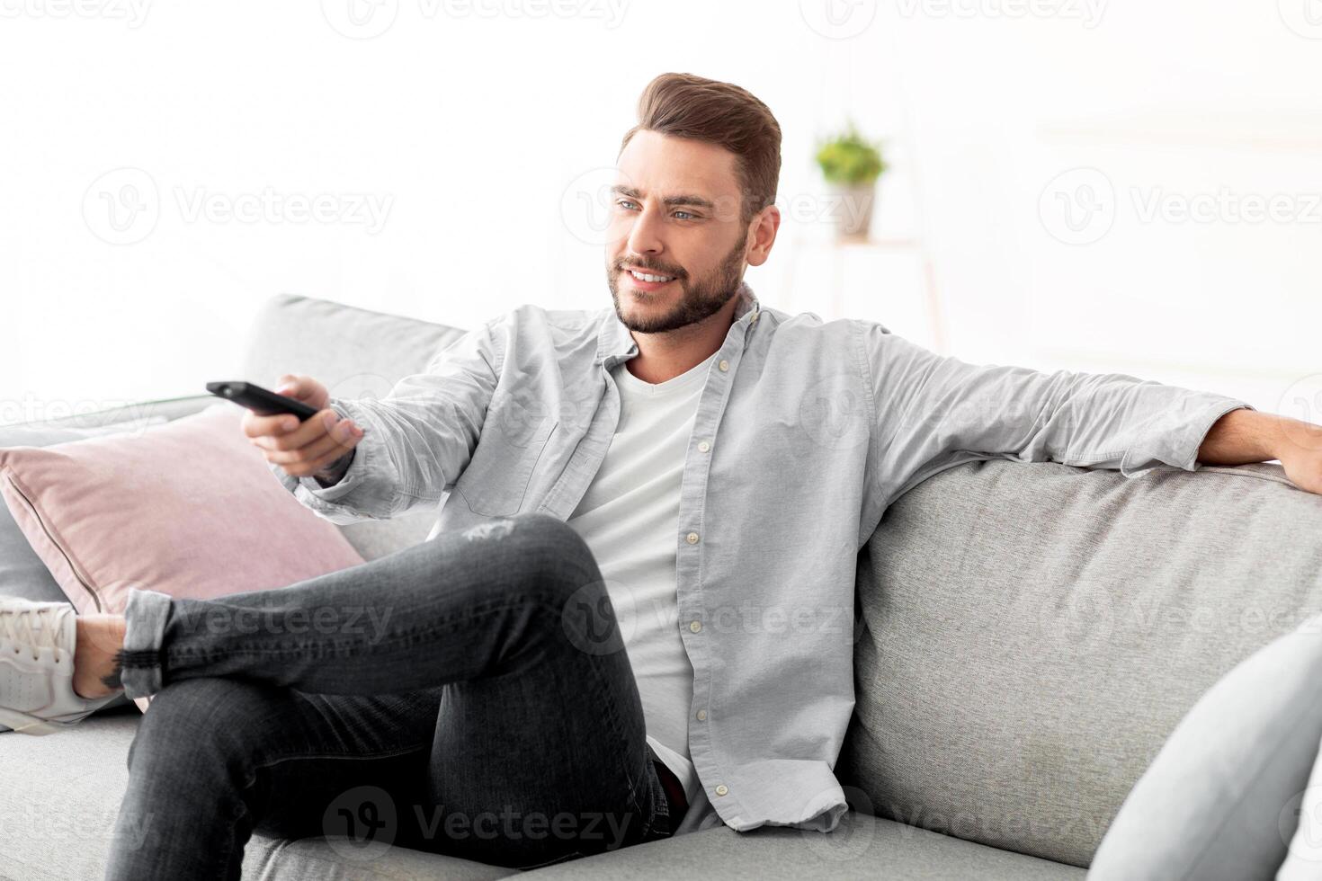 Leisure and entertainment. Happy guy watching TV switching television channels with remote control, sitting on sofa at home, copy space. TV programming concept photo