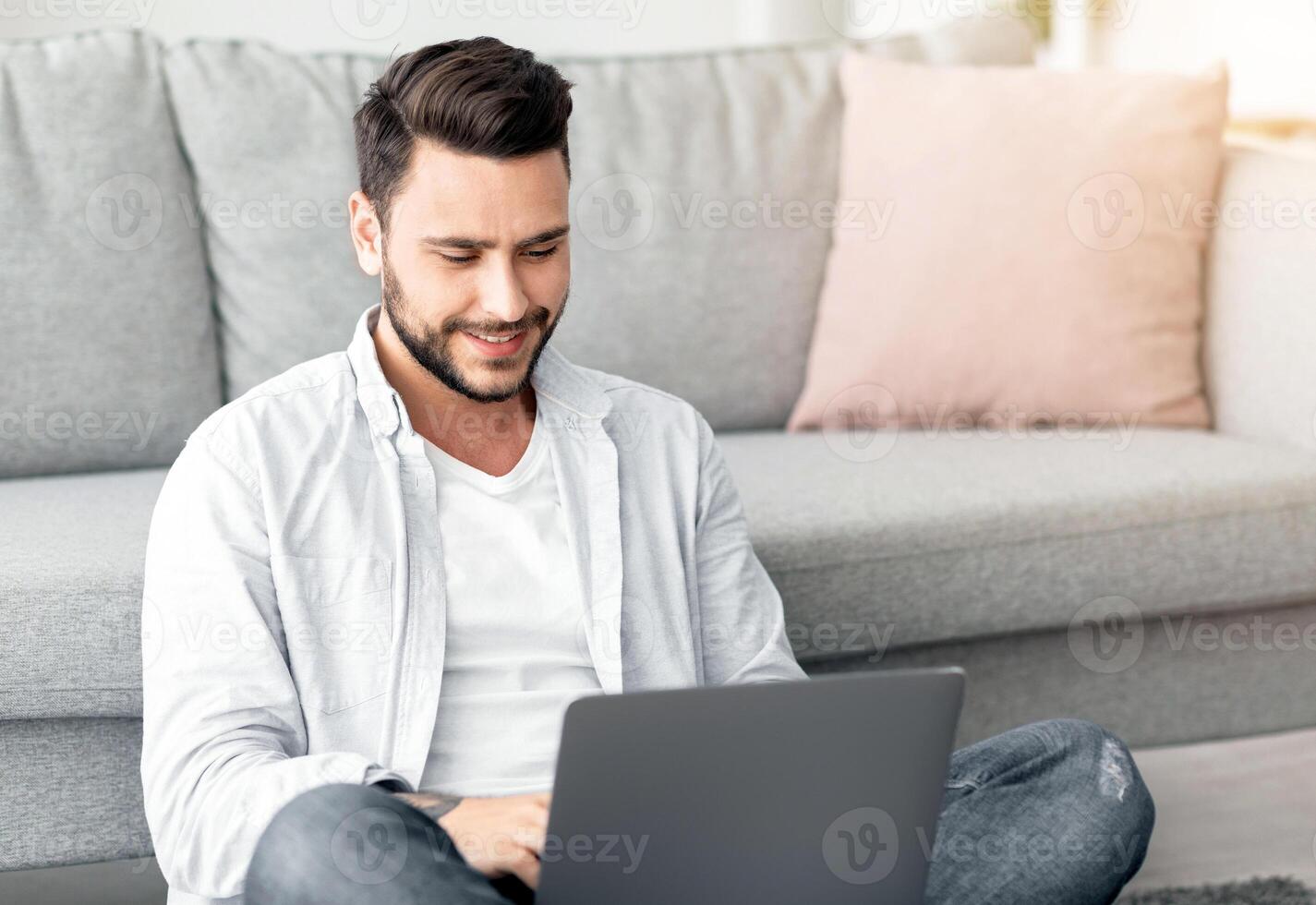 Smiling millennial man working from home, sitting on floor and using laptop, free space. Happy young guy enjoying his weekend, surfing on internet on laptop photo