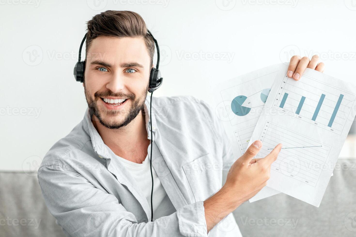 Online meeting concept. Millennial man having call, showing diagrams to laptop camera, discussing financial reports, wearing headset while sitting at home interior photo