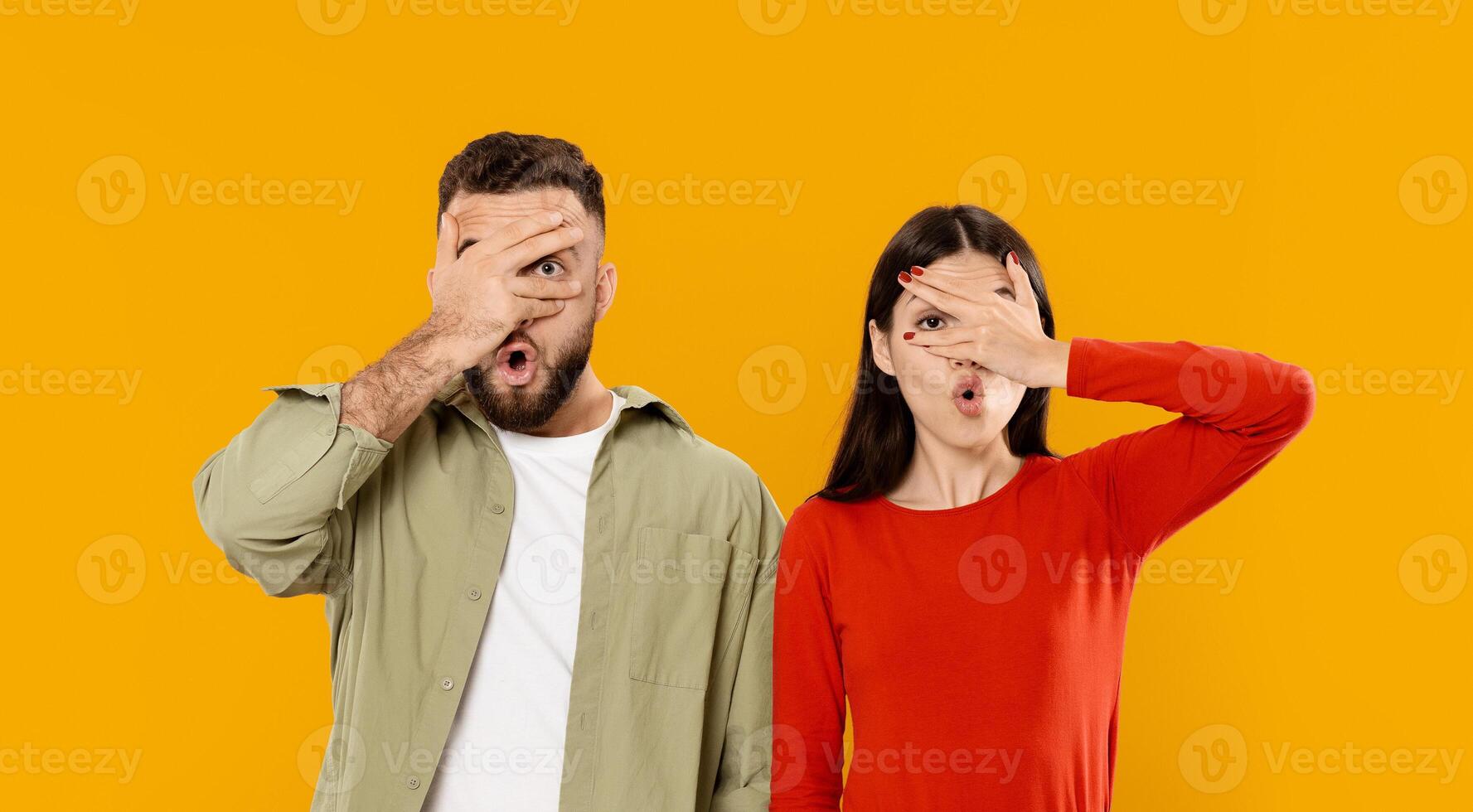 A young Caucasian couple stands together against a bright yellow background. They engage in a playful gesture, covering their faces with their hands while expressing joy. photo