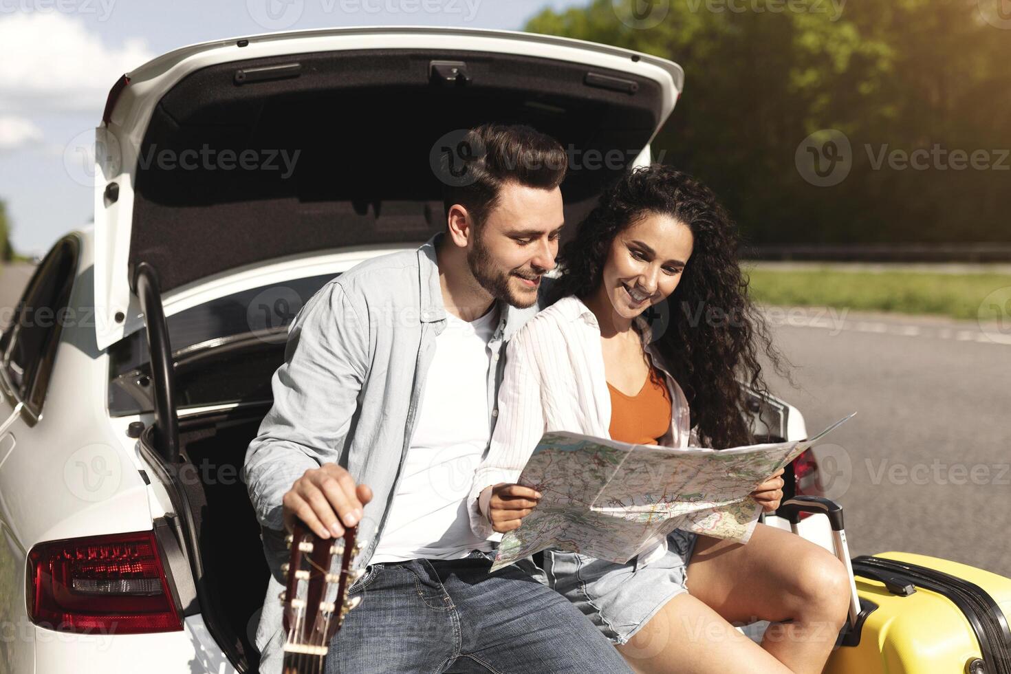 Positive millennial couple going on road trip in car, looking at map, taking break from driving, sitting in auto trunk together. Young Caucasian guy and his girlfriend having summer holiday trip photo