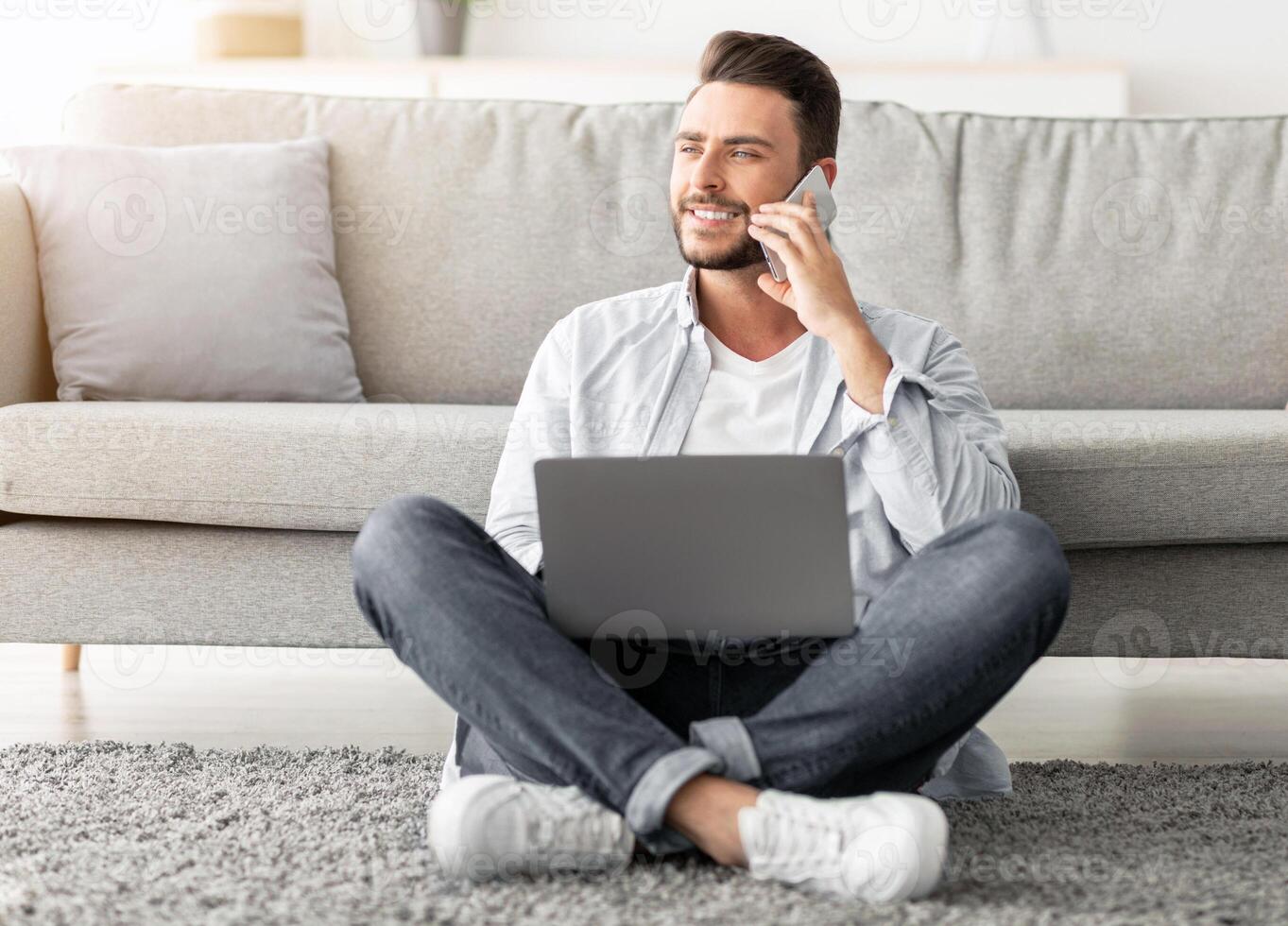 Distant work and mobile communication. Happy freelancer man talking on cellphone and using laptop while working remotely from home, sitting on floor near sofa in living room, free space photo