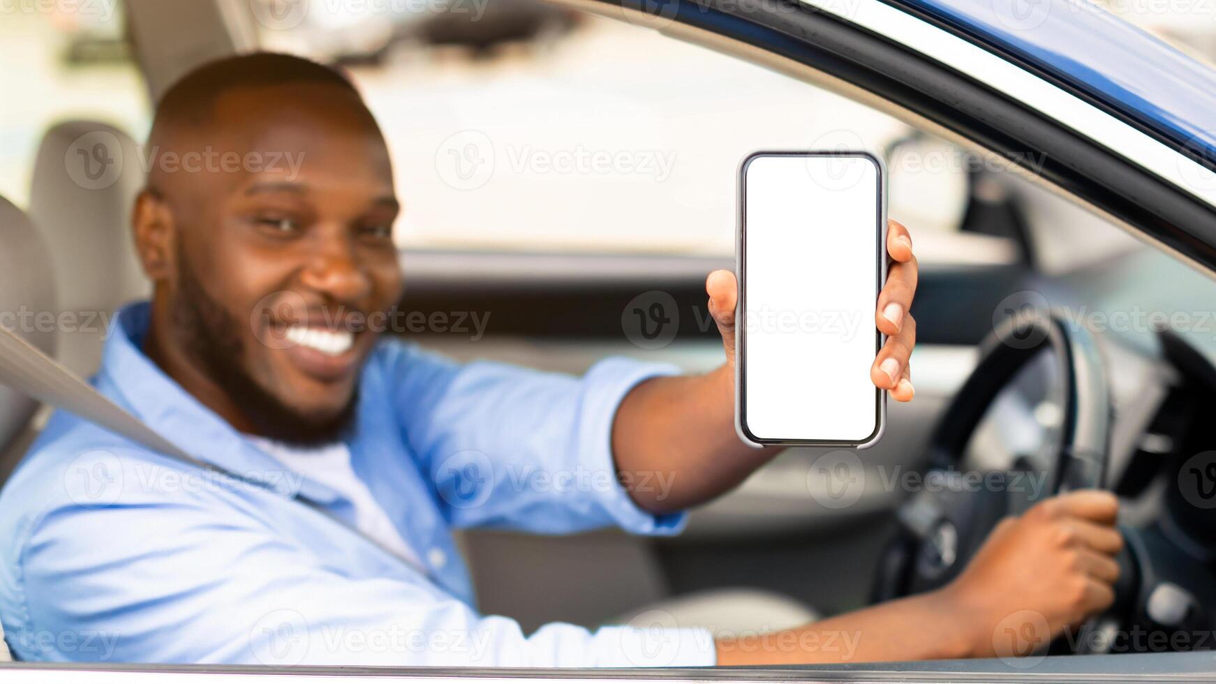 Closeup of young smiling African American man showing mobile phone with white empty screen for mock up sitting on driver seat in new luxury car, holding gadget in hand, selective focus on device photo