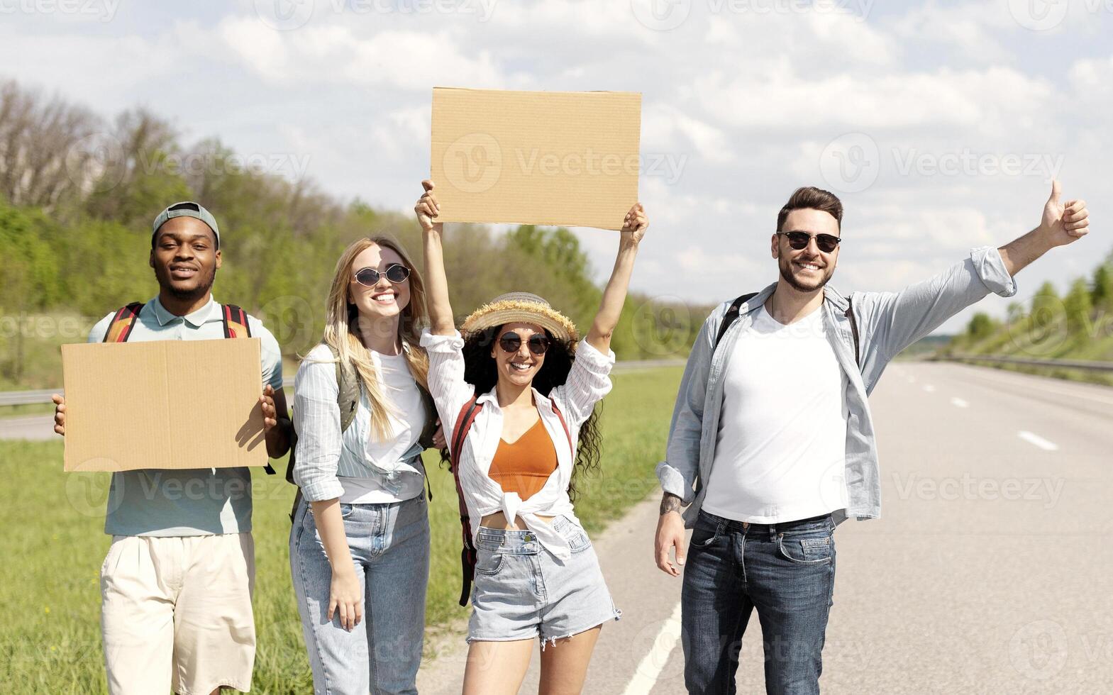 Happy diverse friends standing on highway with empty signs, stopping car, going on trip together, outdoors. Group of multinational hitchhikers traveling by autostop, having fun during road trip photo