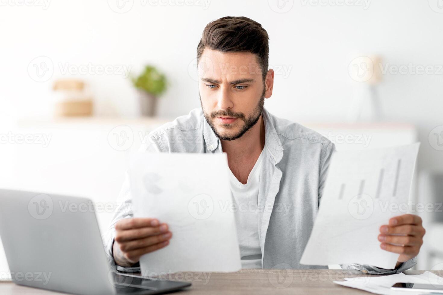 Millennial male self-entrepreneur working with papers at desk at home office, checking annual financial reports, free space. Young man reading documents, sitting at workplace with computer photo