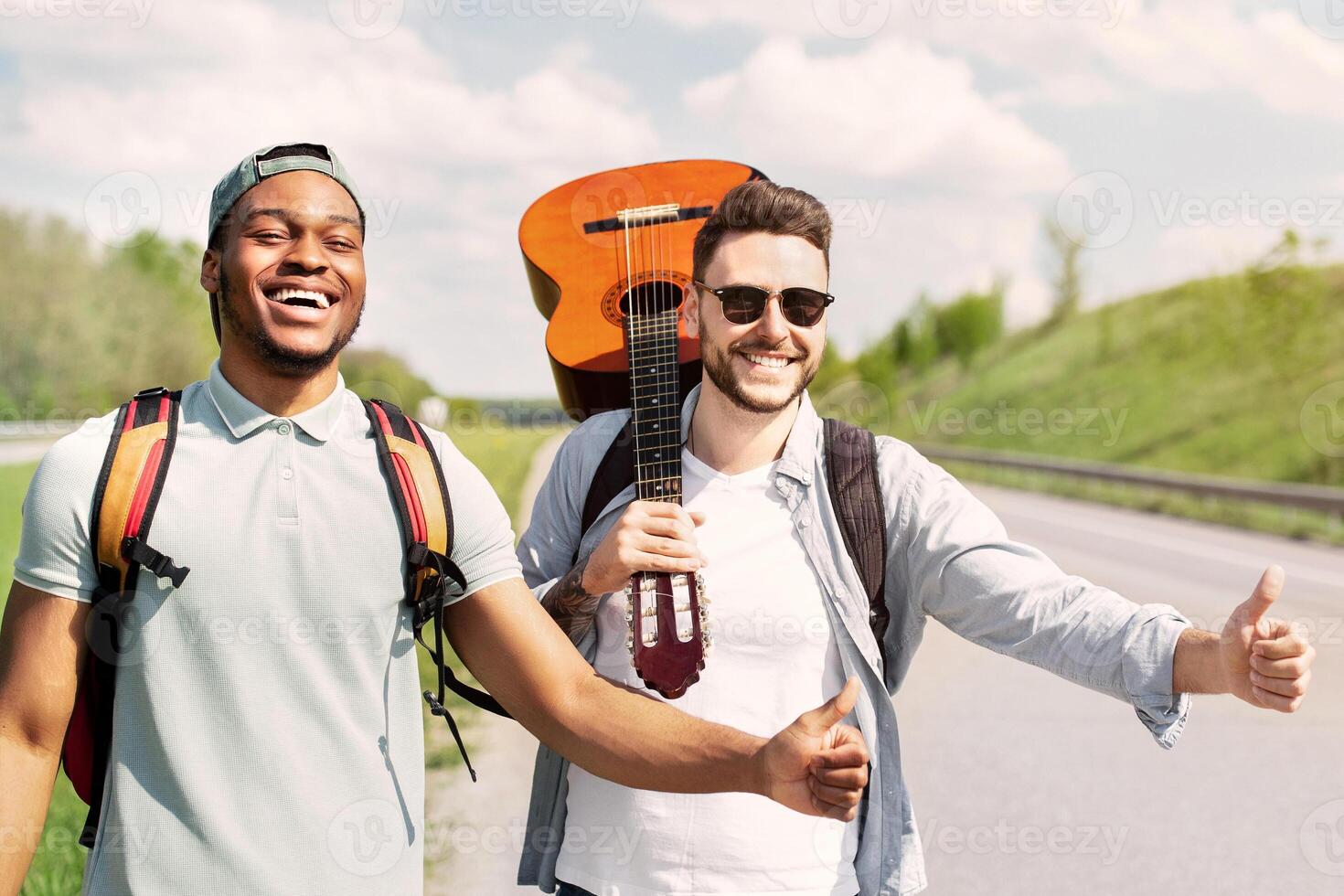 Two guys with guitar hitchhiking on road, stopping passing cars with thumb up gesture, having fun summer adventure, copy space. Multiracial friends traveling by autostop, enjoying their journey photo