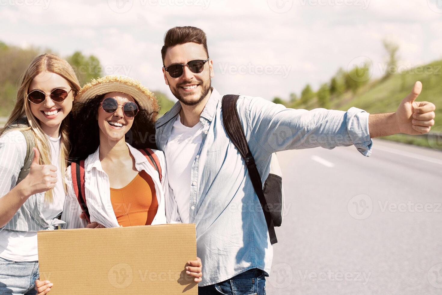 Portrait of happy diverse friends hitchhiking together on highway, making thumb up gestures, holding empty signs with space for mockup. Joyful young people having autostop trip in summer photo