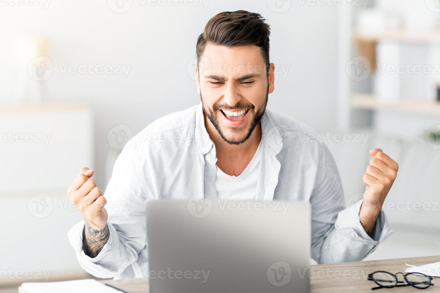 Portrait of overjoyed guy celebrating success, shaking clenched fists while sitting at desk in living room interior, copy space. Man using laptop computer, reading great news, screaming yes photo