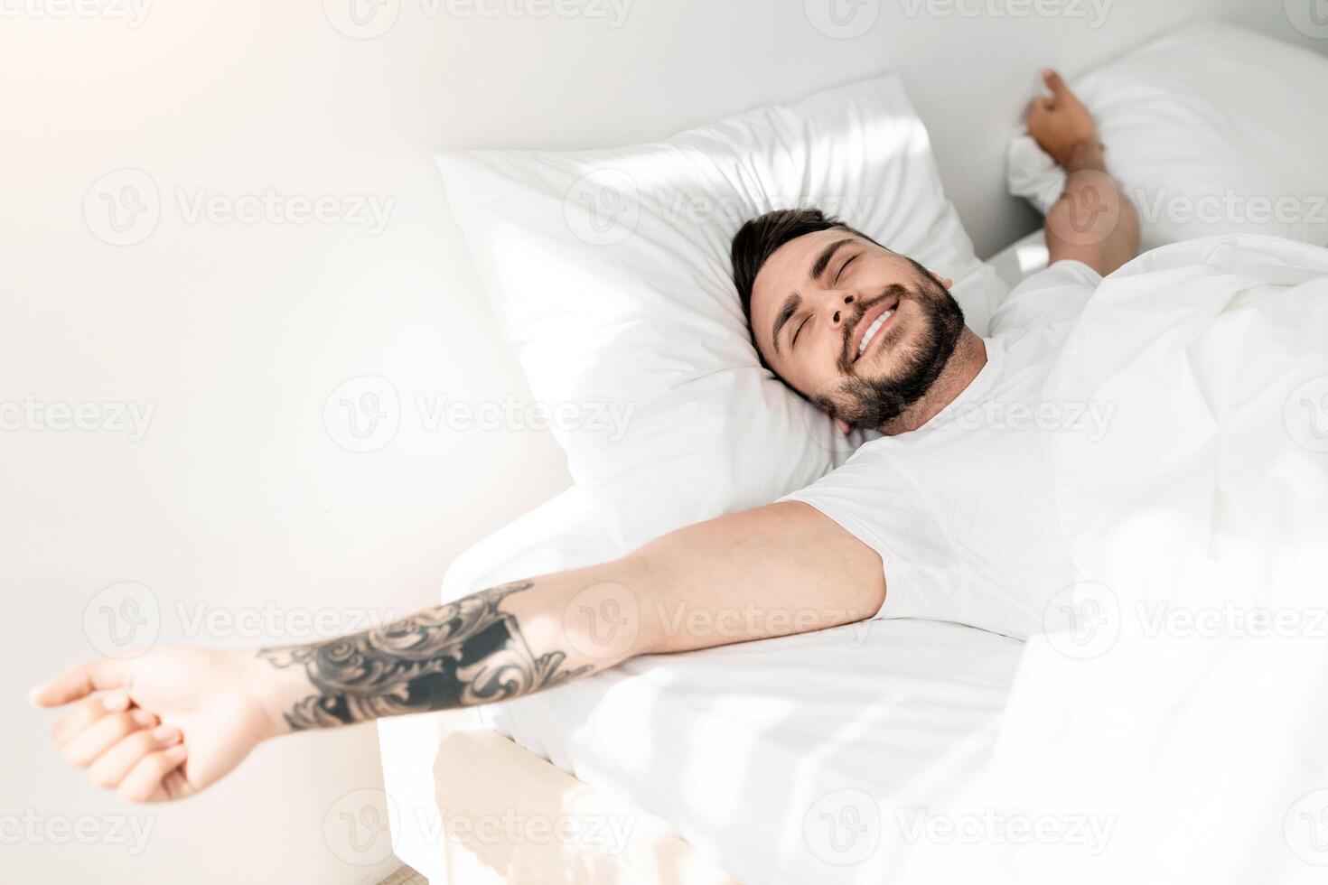 Comfortable sleep. Wellslept tattooed man waking up in morning and stretching hands, smiling with closed eyes while lying in bed, free space. Good morning concept photo