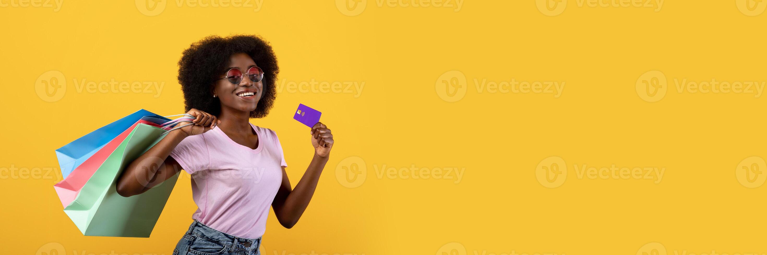 Seasonal sales concept. Happy black woman in sunglasses with colorful shopping bags and unlimited credit card smiling to camera on yellow background, studio shot, copy space photo