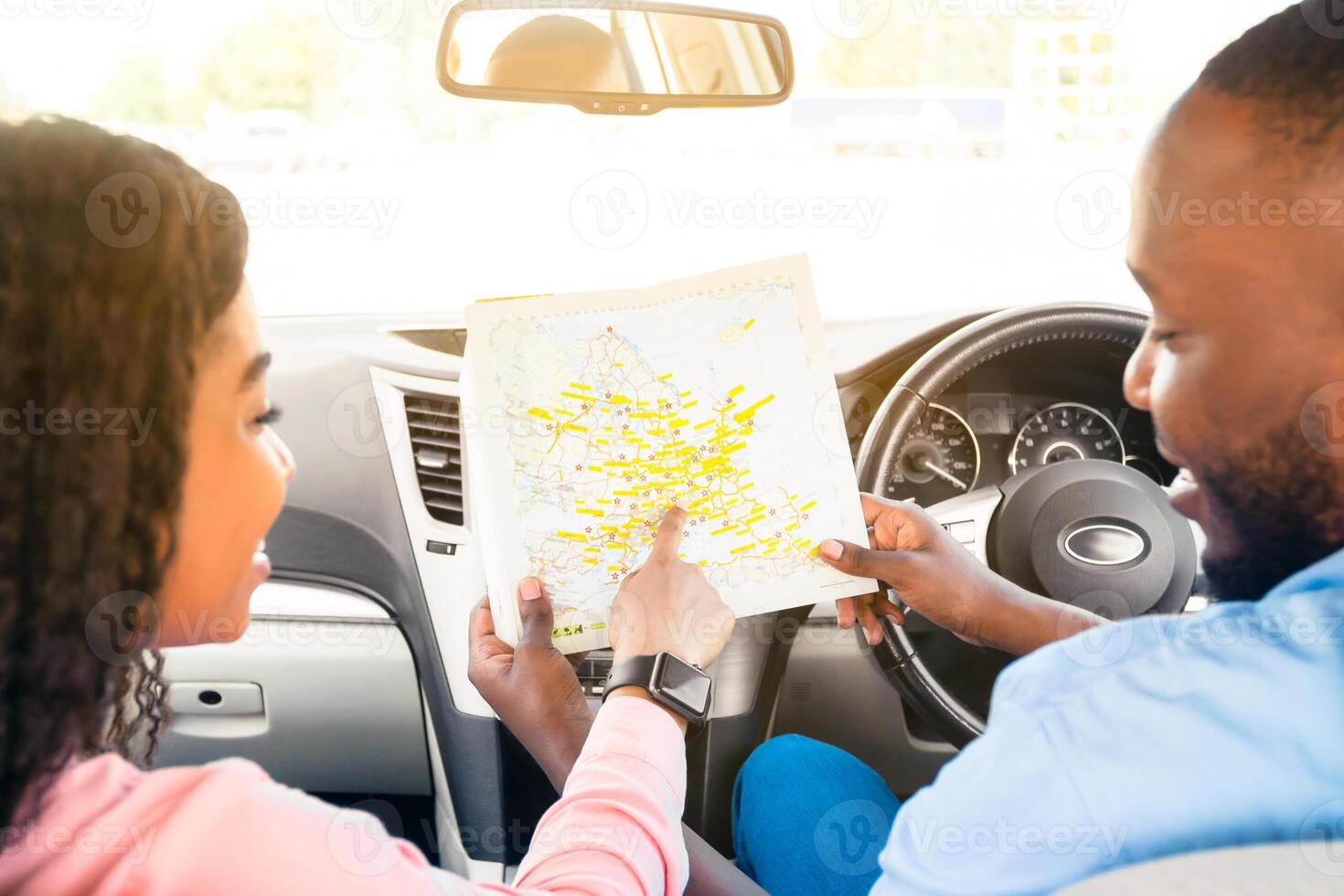Road Trip Concept. Rear back view from passenger's seat of smiling black man and lady holding and looking at paper map sitting inside car, choosing location, woman pointing at destination point photo