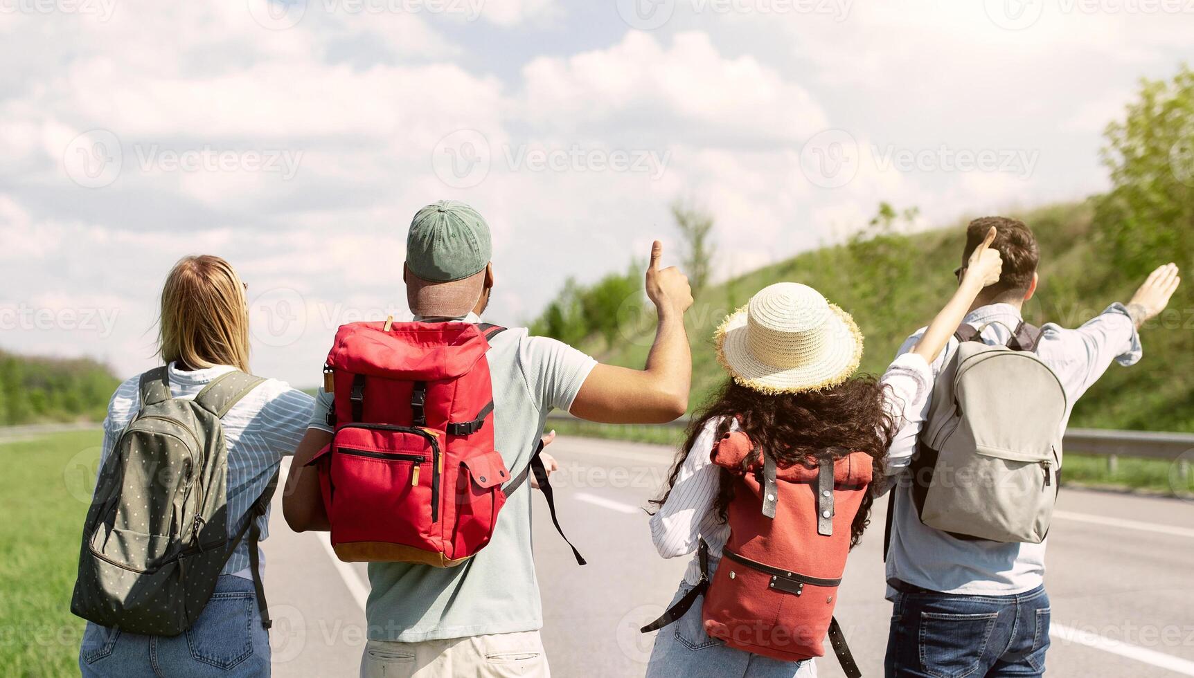 Back view of diverse friends with backpacks traveling by autostop, hitchhiking, trying to get free ride in countryside. Multiethnic young students having summertime adventure, going on road trip photo