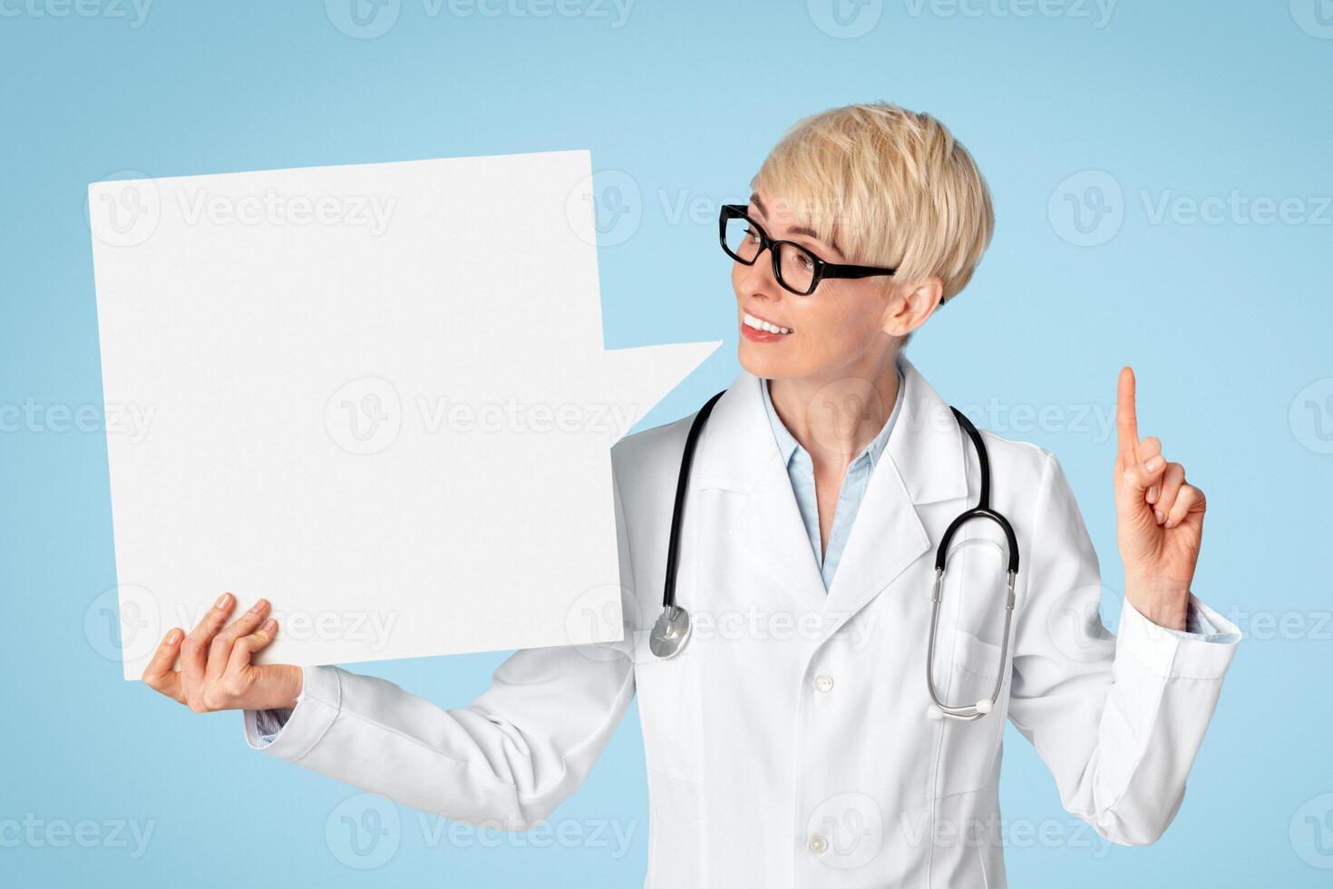 Doctors advice, medicine and prevention illness. Adult female doctor in glasses and white coat looks at abstract space for words and points finger up, isolated on blue background, studio shot photo