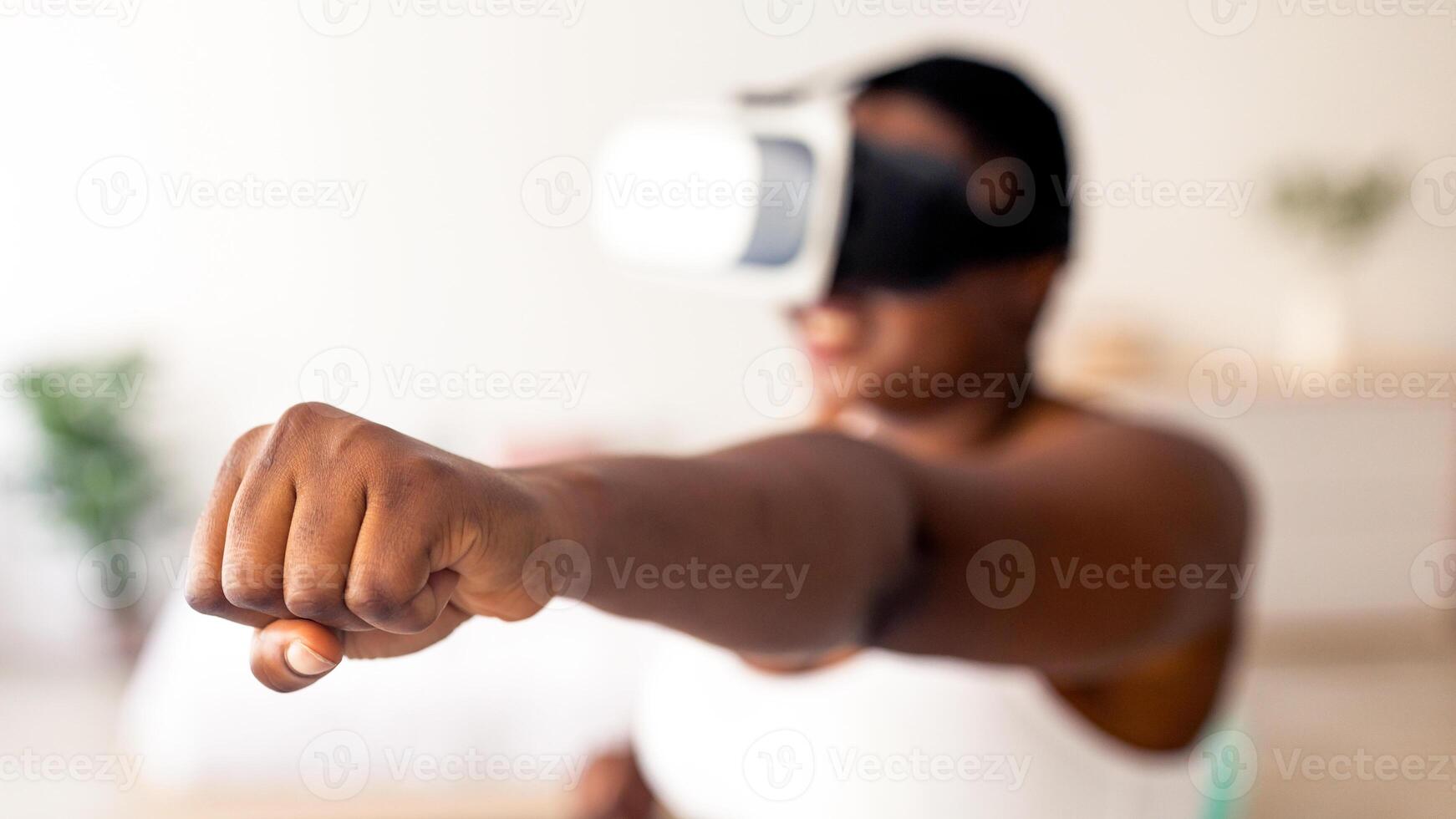 Virtual training concept. Overweight black lady in VR glasses using fighting simulator, making punch at home, focus on clenched fist. Plus size Afro woman boxing with augmented reality app photo