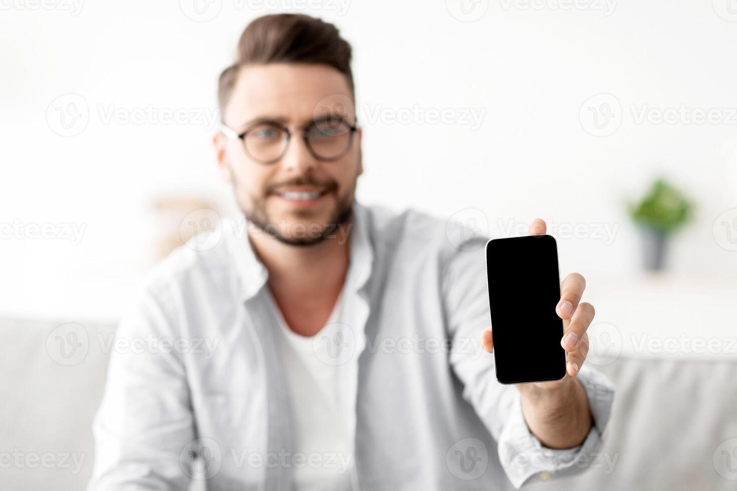 I recommend this website. Young man showing smartphone with blank screen to camera, mockup for your ad or design. Guy sitting on sofa at home, holding cellphone in hand, selective focus photo