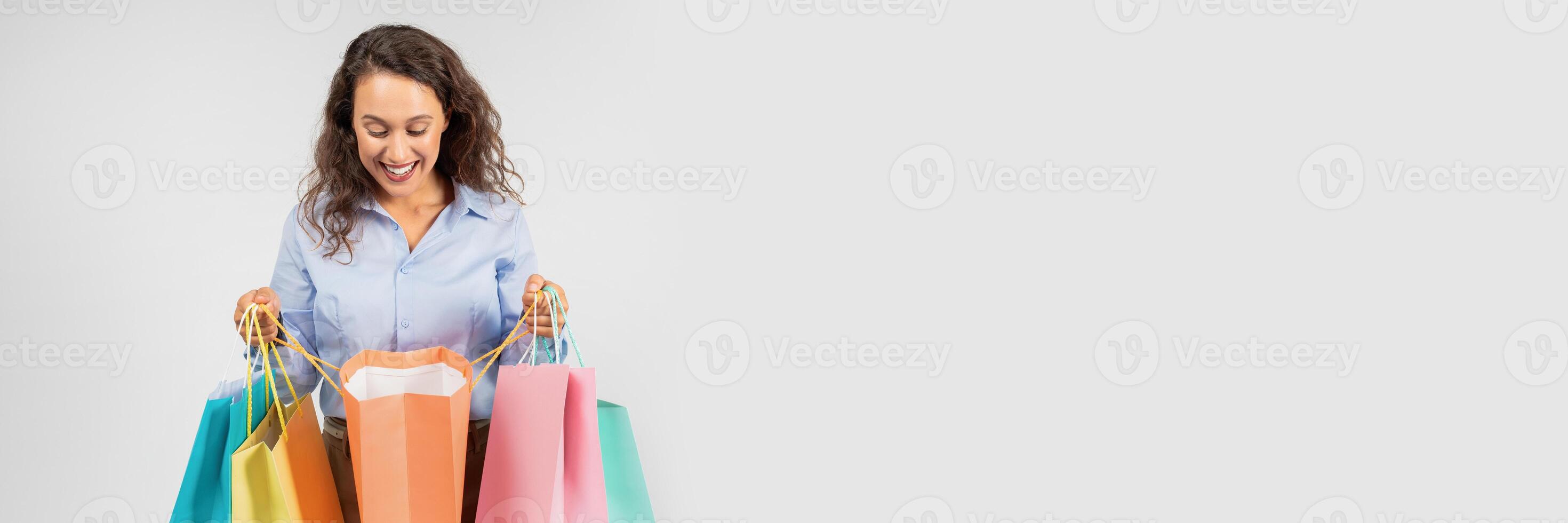 An exuberant young woman with curly hair, clad in a blue shirt, looks down at her multiple bright shopping bags with a delighted smile, enjoying a successful shopping spree, copy space photo