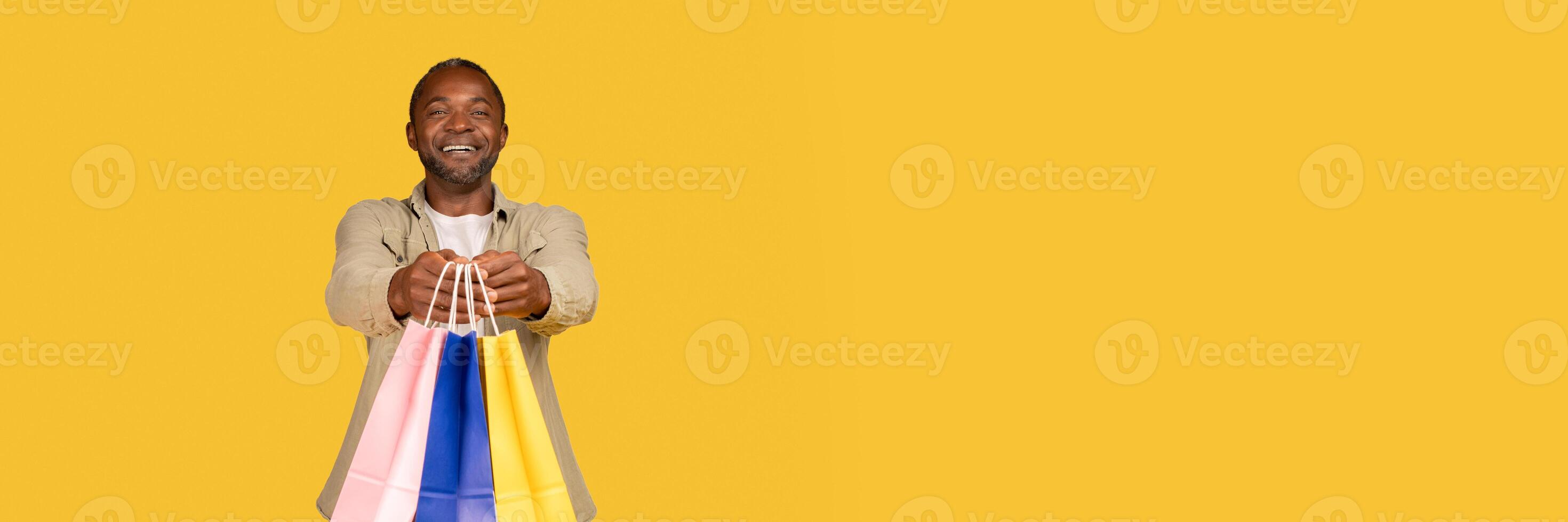 Cheerful african american middle aged man shopaholic show many packages with purchases from shop, recommend buy, isolated on orange background, studio, copy space. Online shopping, delivery and sale photo