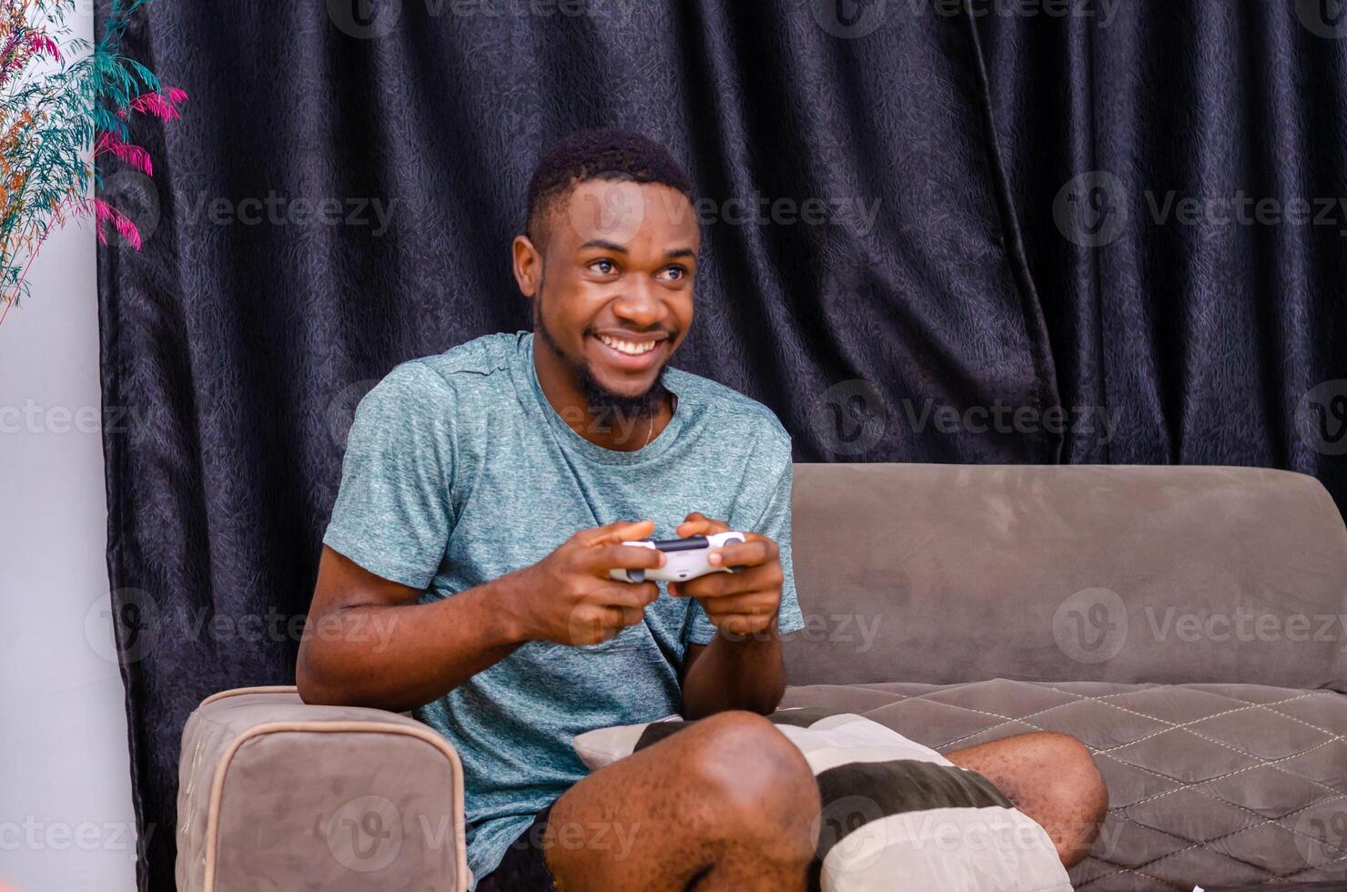 Front view of young bearded hipster man sitting on the couch in the living room staying at home and playing, holding wireless joystick photo