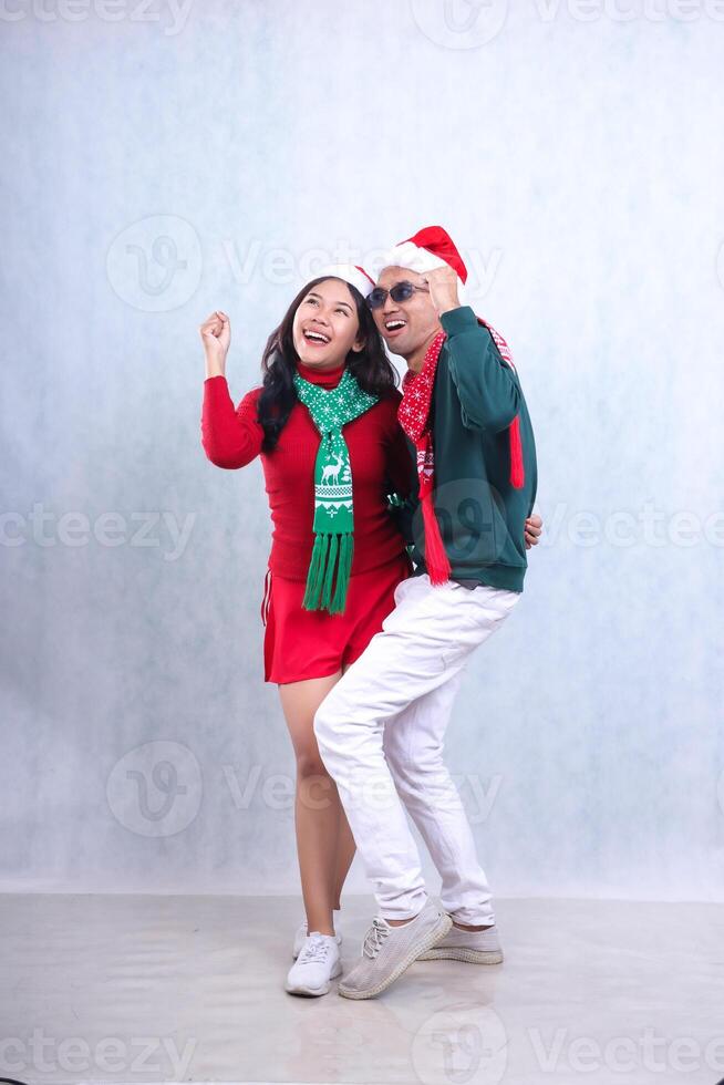 full length Asian couple wearing party clothes with Santa hats celebrating Christmas and New Year, hands raised up in fists look at left upper corner cheerfully. isolated on white background photo