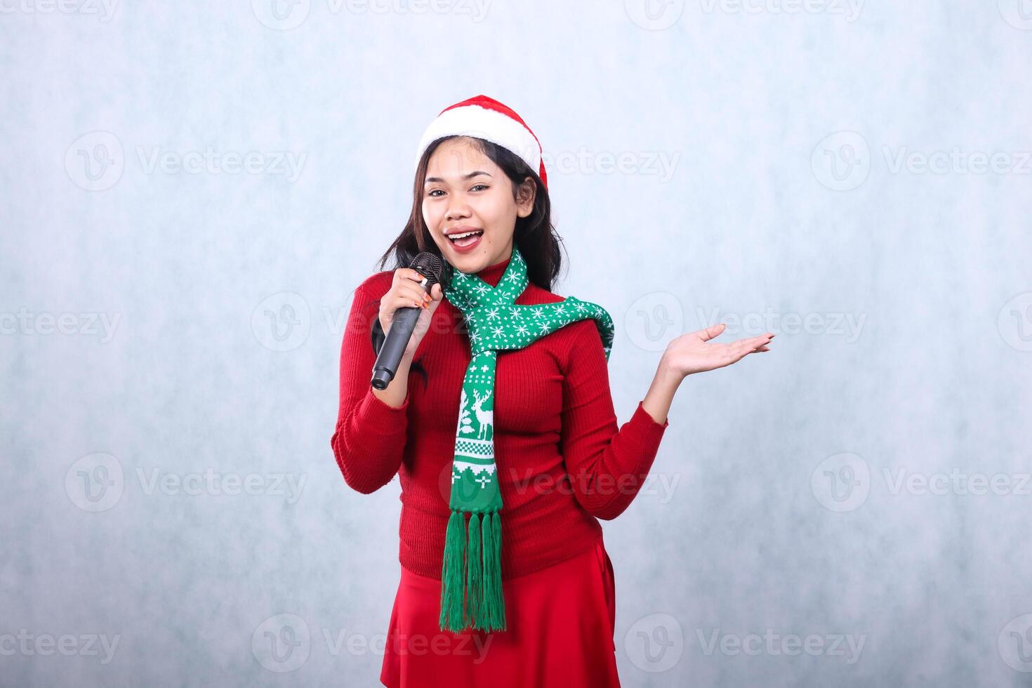 beautiful woman wearing red sweater with Santa hat and scarf celebrating merry christmas, happy singing to camera holding karaoke mic hand presenting to the side, isolated on white background photo