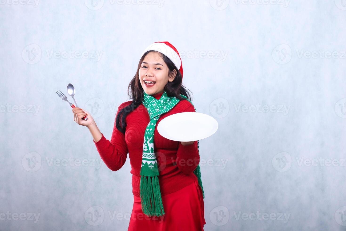 beautiful woman wearing red sweater red sweater with santa hat and scarf celebrating merry christmas, cheerful to camera carrying white plate and holding fork spoon, isolated on white background photo