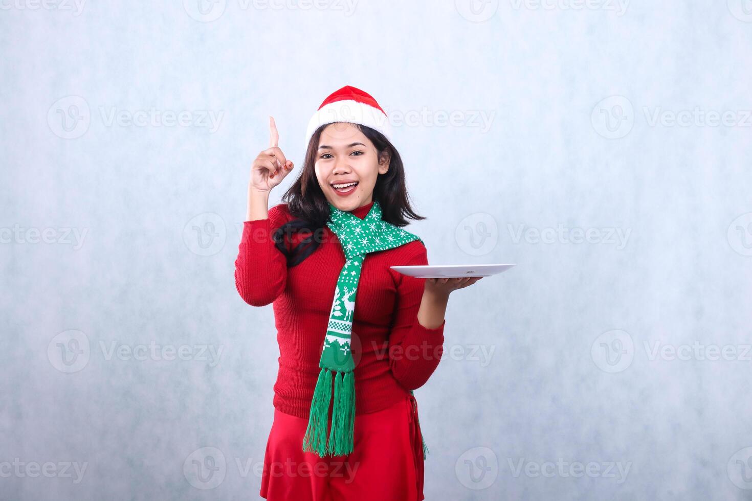charming adult asian woman wearing red sweater with Santa hat and scarf, cheerful to camera holding white plate pointing up, celebrating merry christmas and happy new year isolated on white background photo