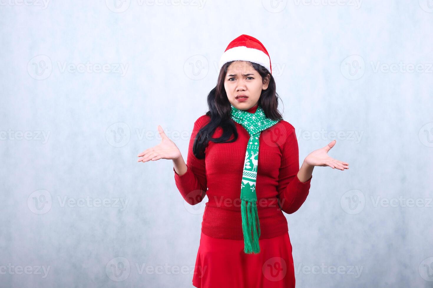 Asian woman expression wearing red sweater party wear with Santa hat and scarf celebrating christmas and new year, both hands open to the side surprised hesitant, isolated on white background photo
