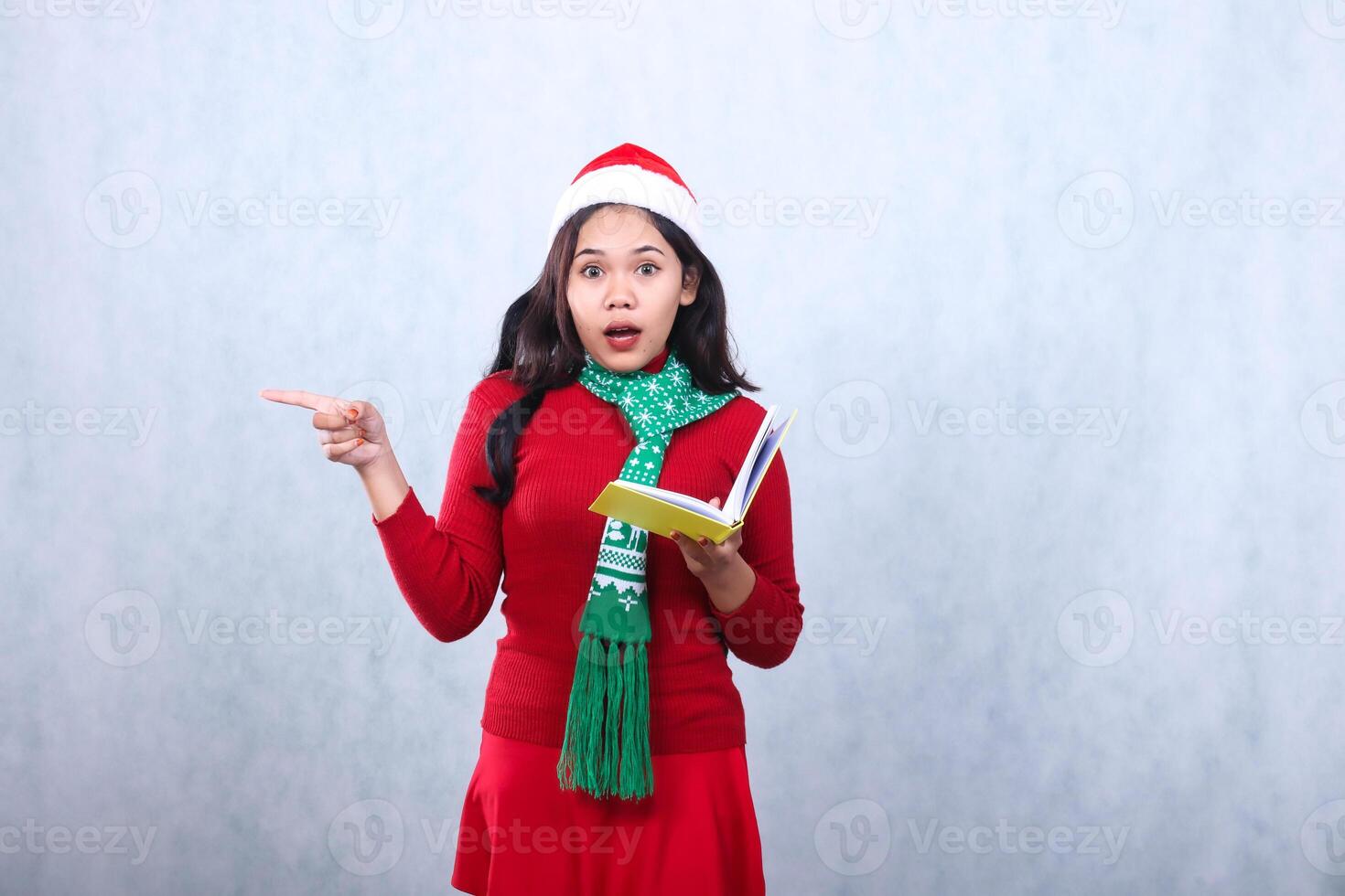 Charming young woman wearing red sweater red sweater with Santa hat and scarf, surprised to camera opening book while pointing to right, celebrating merry christmas isolated on white background photo