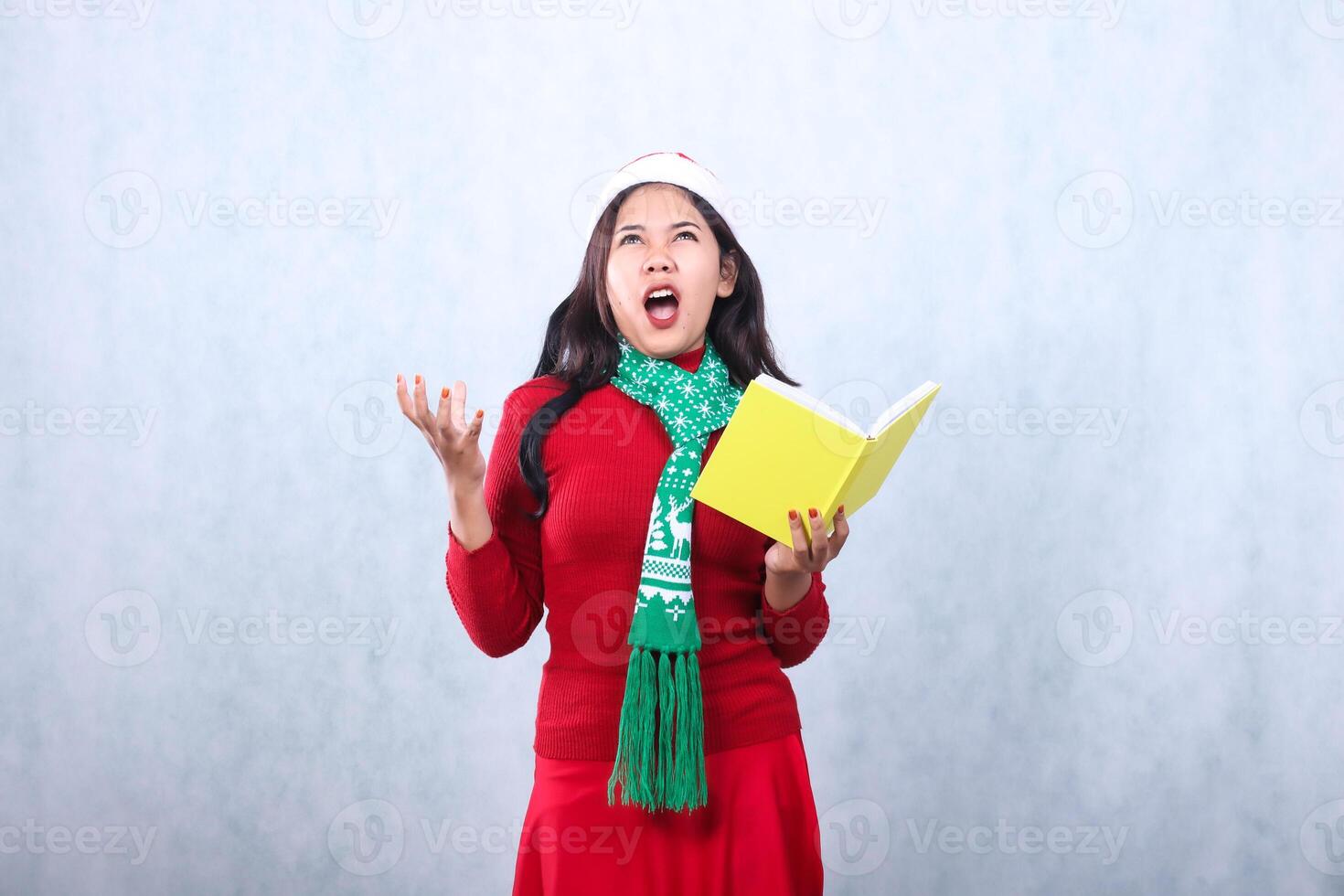 expression of young woman wearing red sweater red sweater with santa hat and scarf celebrating merry christmas, screaming up holding book in hand growling stressed, isolated on white background photo