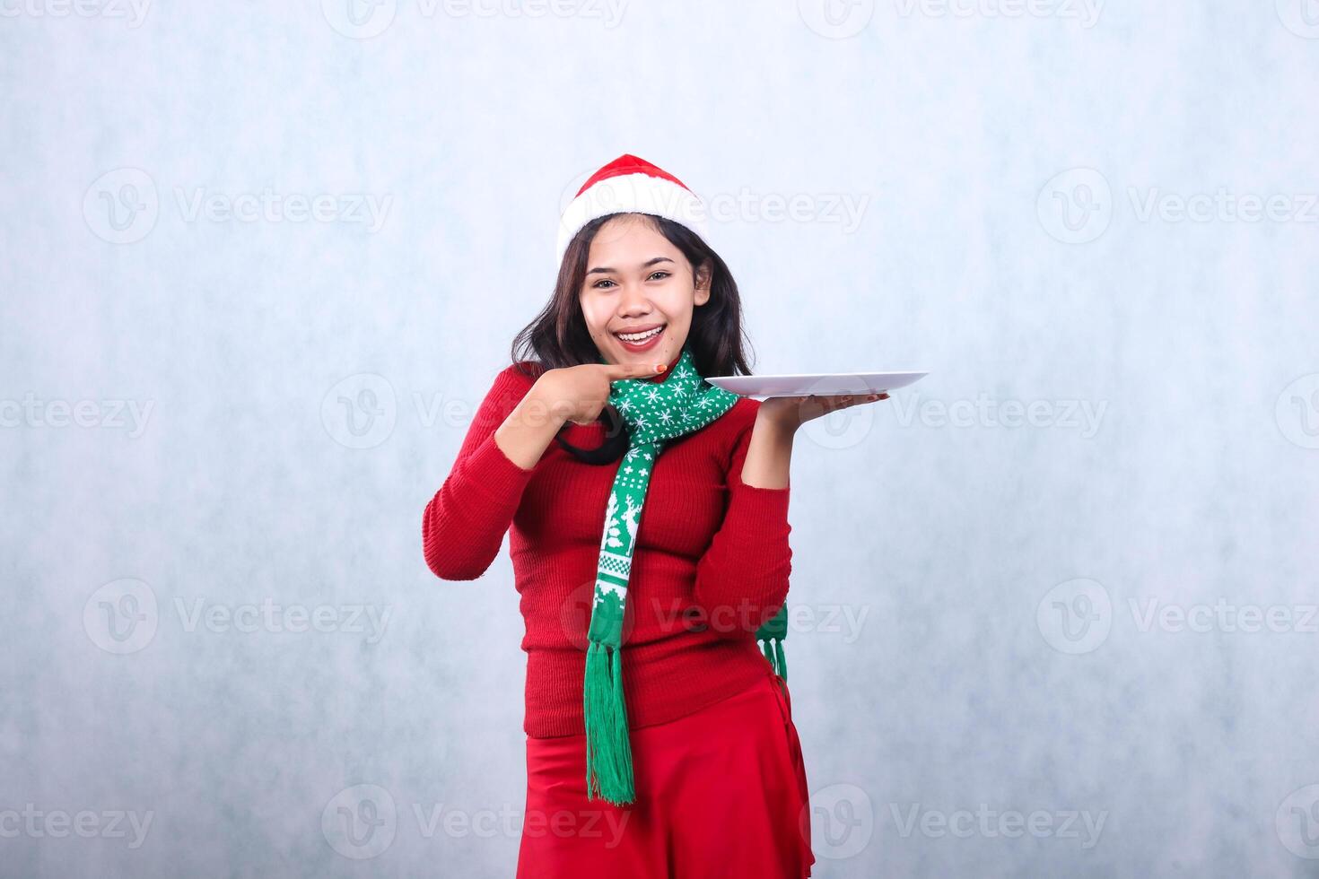 gesture woman wearing red sweater with Santa hat and scarf, cheerful looking at camera hand lifting white plate and pointing at it, celebrating merry christmas isolated on white background photo