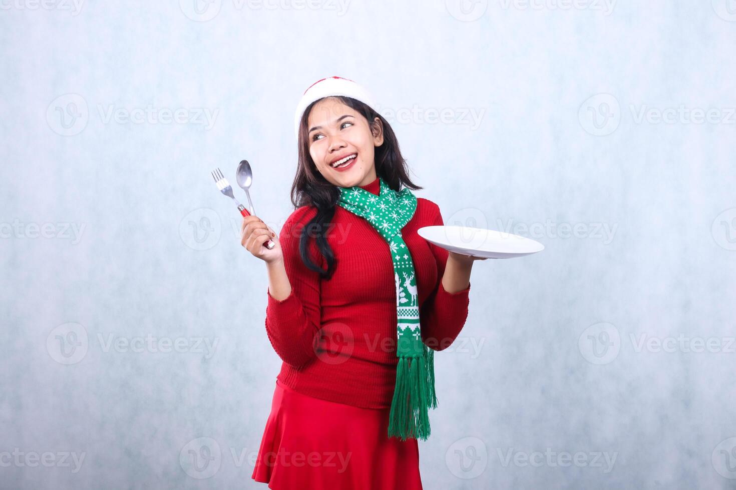 portrait of girl wearing red sweater red sweater with santa hat and scarf celebrating merry christmas, cheerful to the right carry white plate and hold spoon fork, isolated on white background photo