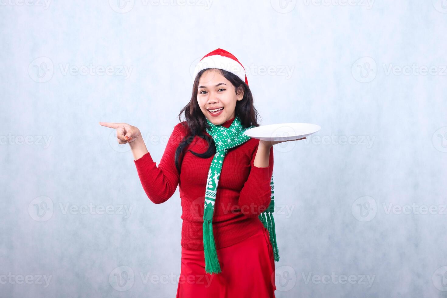 young girl wearing red sweater with Santa hat and scarf, cheerful looking at camera, hand lifting white plate and pointing to the right, celebrating merry christmas isolated on white background photo