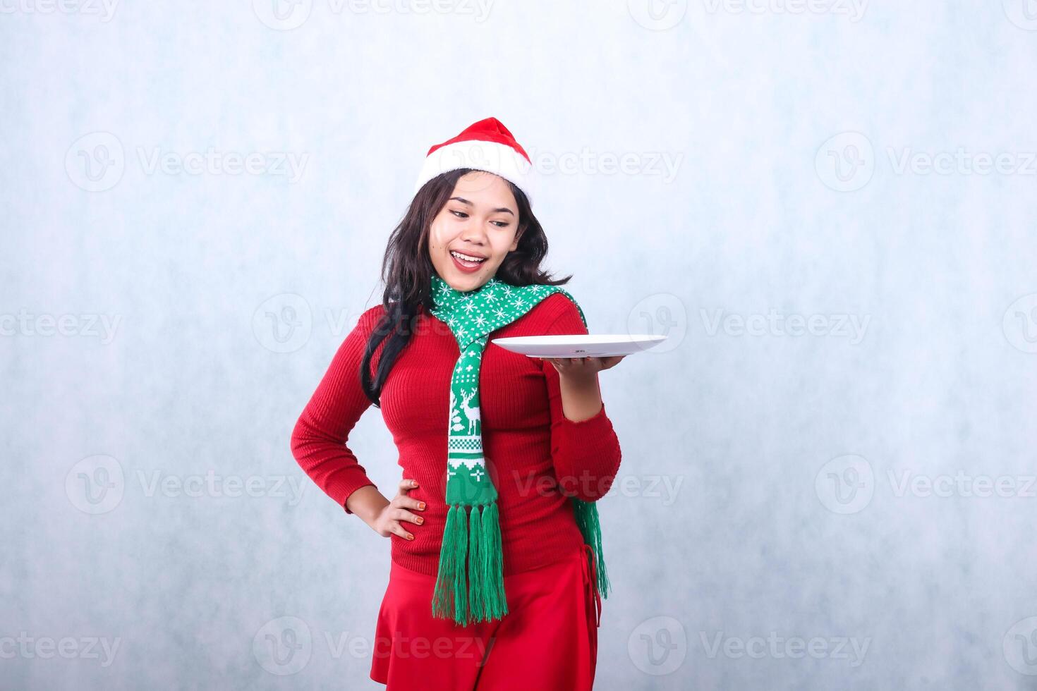 Charming young indonesia woman wearing red sweater with Santa hat and scarf, cheerful to the plate holding white plate and waist, celebrating merry christmas and new year isolated on white background photo