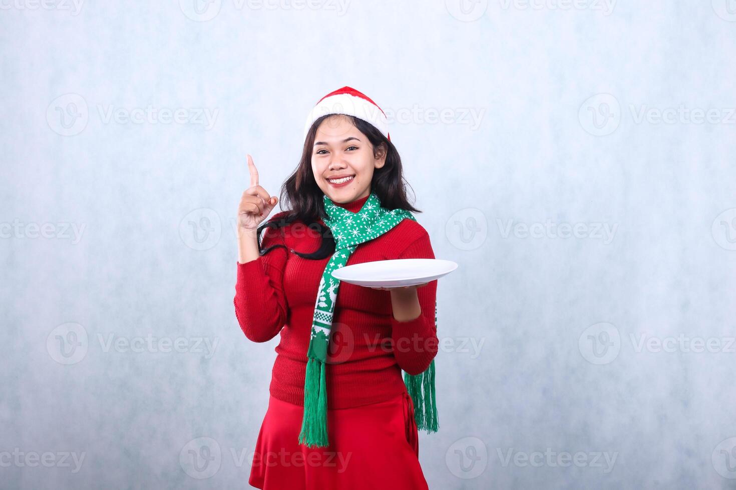 Charming young woman wearing red sweater with Santa hat and scarf, happy looking at camera, hand holding white plate and pointing at it, celebrating merry christmas isolated on white background photo