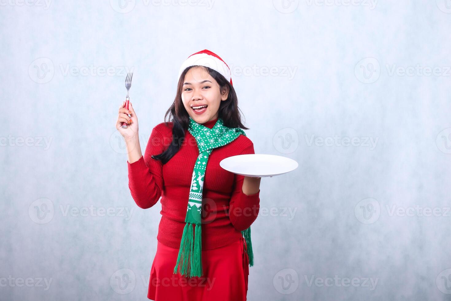 Charming Asian woman wearing red sweater with Santa hat and scarf, cheerful to camera carrying white plate and holding fork aside, celebrating merry christmas and new year isolated on white background photo